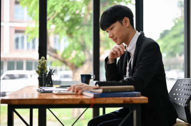 Arkadan bakıldığında, resmi bir iş elbisesi içinde mutlu ve zeki bir Asyalı işadamı masasındaki bir belgeyi okumaya odaklanır..