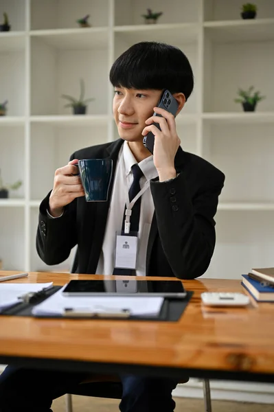彼の机の上で彼のクライアントと電話で話している間 正式なビジネススーツでプロとスマート若いアジアのビジネスマンや男性銀行家はコーヒーを飲んでいます — ストック写真