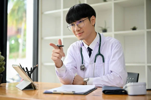 一位穿着制服的英俊而聪明的千年亚洲男医生坐在办公桌前 用手指对着摄像机 — 图库照片
