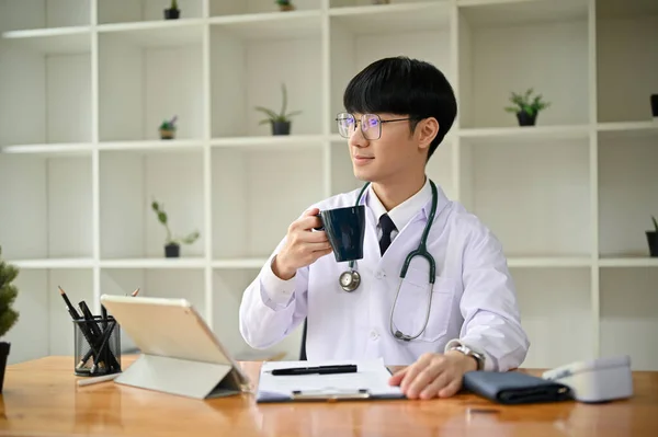 一位身穿白袍 聪明而专业的千年期亚洲男医生 一边朝外张望 一边做着白日梦 一边在办公室的办公桌边喝着早茶 — 图库照片