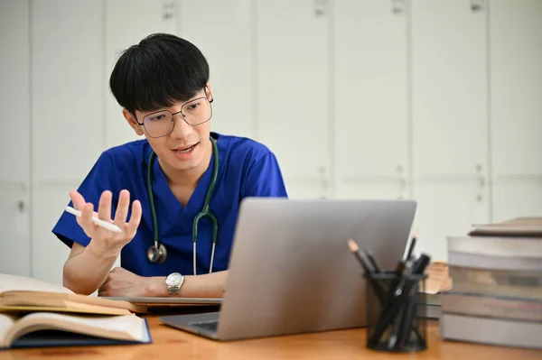 一位聪明而专业的年轻亚洲男性医科学生或医生正在和他的医疗团队举行在线会议 或者在他的房间里与一位病人进行在线体检 — 图库照片