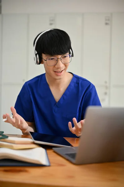 A focused Asian male medical student or doctor in scrubs wearing headphones is having an online medical checkup with a patient or joining a medical webinar. Medical and technology concept