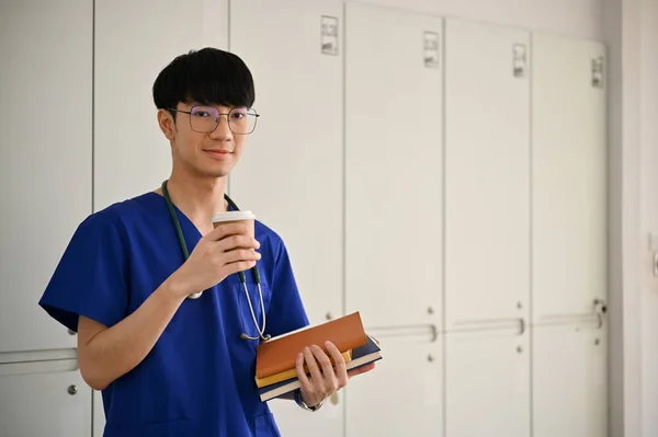 在医院的走廊里 站着一个英俊而积极的亚洲年轻医学专业学生或医生的画像 手里拿着咖啡杯和书本 — 图库照片