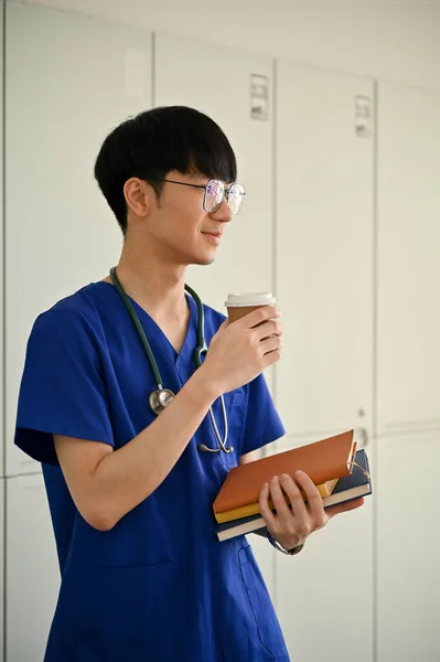 在医院走廊里 一个英俊的年轻的亚洲医学专业学生或擦拭医生的画像 手里拿着咖啡杯和书本 保健概念 — 图库照片