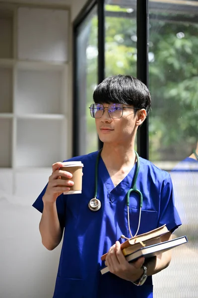 站在医院走廊里的一位英俊聪明的亚洲年轻医学专业学生或医生 手持书本和咖啡杯 带着听诊器 — 图库照片