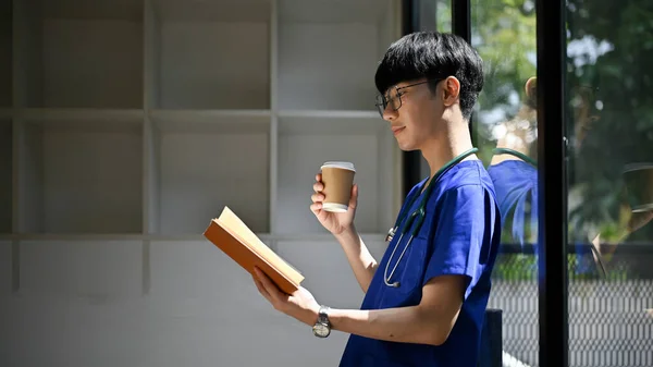 一个英俊而积极的亚洲年轻医生或医科学生靠在玻璃墙上 一边喝着咖啡 一边在办公室看书的背影 — 图库照片