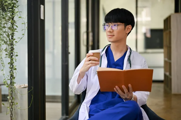 一位身穿制服的英俊而职业的千年期亚洲男性医生 一边看书 一边在办公室喝咖啡 一边朝外张望 幻想着自己在事业上的成功 — 图库照片