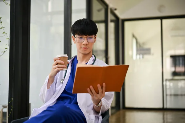 一位聪明而专注的身穿制服的千年期亚洲男医生在医院的办公室看书 喝咖啡 — 图库照片