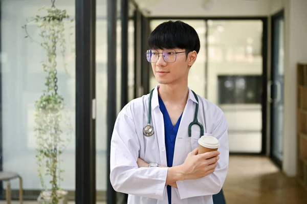一名身穿制服 手持听诊器的英俊而积极的千年亚洲男医生站在医院走廊里 手里拿着咖啡杯 医务工作者概念 — 图库照片