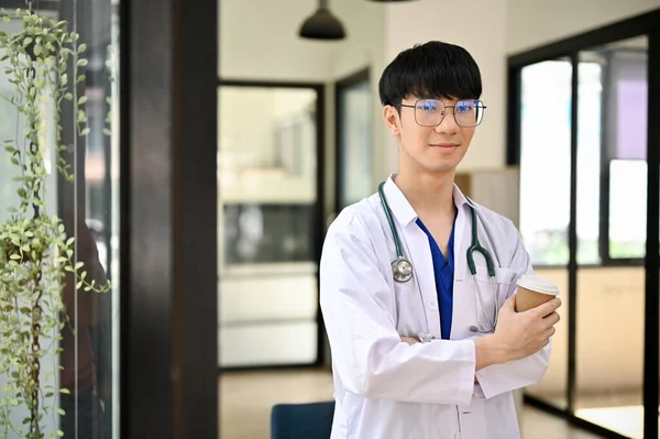 一名身穿制服 手持听诊器的职业而积极的千年期亚洲男医生站在医院走廊里 手里拿着咖啡杯 保健概念 — 图库照片