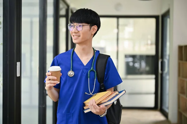 一位快乐而英俊的年轻亚洲医学专业学生或擦拭医生手拿着咖啡杯和书本沿着医院走廊走着 — 图库照片