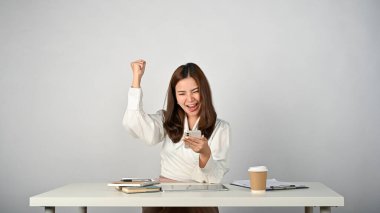 Aşırı mutlu Asyalı genç bir iş kadını masasındaki akıllı telefon ekranına bakarken yumruğunu gösteriyor. Beklenmedik haberler, iyi haberler, sıcak anlaşma, sevinç, kazanma, zafer jesti