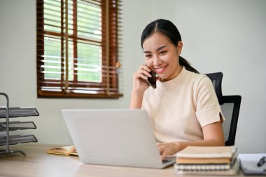 Genç Asyalı kadın, ofisteki bilgisayarını kullanırken iş arkadaşıyla telefonda konuşuyor.. 