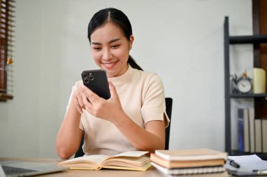 Genç ve çekici Asyalı kadın evinde masasında otururken akıllı telefonunu kullanıyor.. 