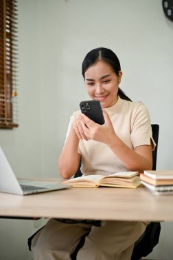 Genç ve çekici Asyalı kadın evinde masasında otururken akıllı telefonunu kullanıyor.. 