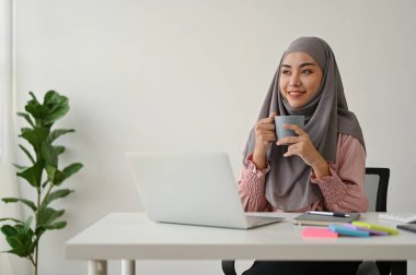Genç, çekici Müslüman iş kadını ofis odasında dizüstü bilgisayarla çalışırken kahve içiyor.. 