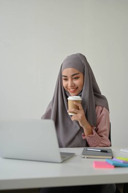 Genç, çekici Müslüman iş kadını ofis odasında dizüstü bilgisayarla çalışırken kahve içiyor.. 