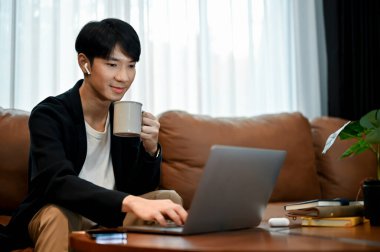 Genç yakışıklı Asyalı adam kanepesinde dinlenirken dizüstü bilgisayar kullanıyor ve boş zamanlarında kahve içiyor.. 