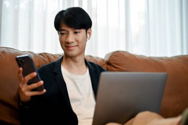 Rahat genç Asyalı adam kanepelerinde oturuyor ve boş zamanlarında elinde akıllı telefon tutuyor.. 