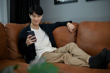 Rahat genç Asyalı adam kanepelerinde oturuyor ve boş zamanlarında elinde akıllı telefon tutuyor.. 