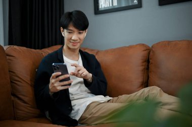 Asyalı genç bir adam kanepelerinde oturup boş zamanlarında elinde akıllı telefon tutuyor. İnsanlar ve teknoloji kavramı