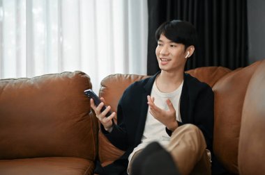 Rahat genç Asyalı adam kanepelerinde oturuyor ve boş zamanlarında elinde akıllı telefon tutuyor.. 