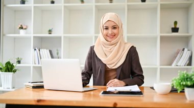 Güzel ve gülümseyen bir Asyalı Müslüman iş kadını ya da kadın patronun portresi modern bir özel ofiste masasında oturuyor..
