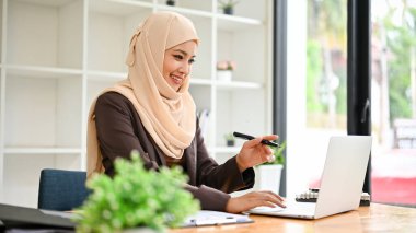 Profesyonel ve çekici Asyalı, tesettürlü Müslüman bir iş kadını bilgisayarındaki iş görevlerine odaklanıyor, ofisinde çalışıyor..