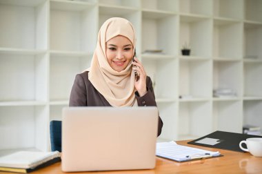 Profesyonel bir Asyalı Müslüman iş kadını iş müşterisiyle telefonda konuşurken masasındaki bilgisayar ekranında bazı detaylara bakıyor..