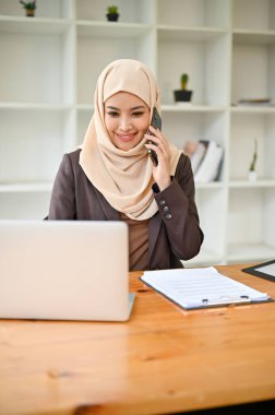 Güzel ve profesyonel bir Asyalı Müslüman iş kadınının portresi bir iş adamıyla telefonda konuşurken masasındaki dizüstü bilgisayarında bazı detaylara bakıyor..
