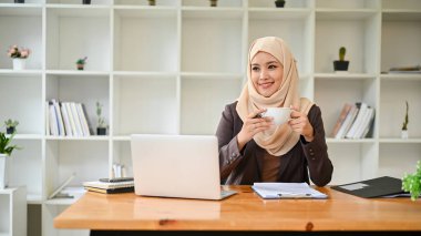 Güzel ve gülümseyen Asyalı tesettürlü Müslüman bir iş kadını ofisteki masasında kahvesinin keyfini çıkarırken mutluluk ve hayallerle pencereden dışarı bakıyor..