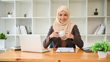 Güzel ve başarılı bir Asyalı Müslüman iş kadını ya da tesettürlü kadın bir patronun portresi. Elinde kahve fincanıyla masasında otururken gülümseyip kameraya bakıyor..