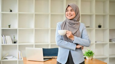 Kendine güvenen ve gülümseyen Asyalı Müslüman bir iş kadınının portresi modern ofisinde elinde kahve fincanıyla duruyor..