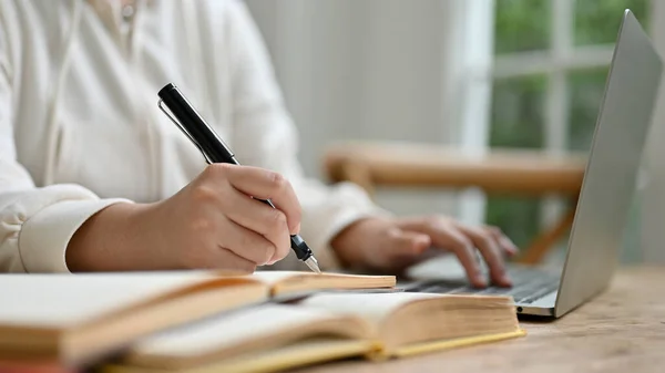 stock image Close-up image of a female college student in a comfy sweater holding pen, writing or taking notes on a paper, doing homework, and using her laptop while sitting in a coffee shop.