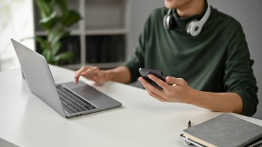 Ofisindeki masasında akıllı telefonunu ve bilgisayarını kullanan nedensel giysiler içinde genç bir Asyalı 'nın yakın plan fotoğrafı..