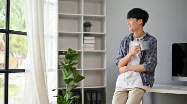 Flanel gömlekli mutlu ve yakışıklı bir Asyalı erkek, elinde kahve fincanıyla ofisinde duruyor, pencereden dışarı bakıyor ve kariyer başarısını hayal ediyor..