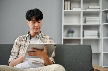 Yakışıklı ve zeki Asyalı bir adam, günlük kıyafetleriyle dijital tabletini kullanıyor. Oturma odasındaki kanepede otururken tablet üzerindeki işine odaklanıyor..