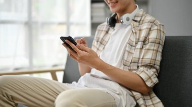 Yakından bakınca, sıradan kıyafetler giymiş yakışıklı bir Asyalı 'nın oturma odasındaki kanepede akıllı telefonunu kullanmasının görüntüsü..