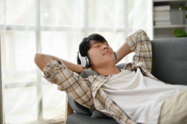 Mutlu ve rahat bir Asyalı adam, oturma odasındaki kanepede uzanmış kulaklıklarıyla müzik dinlemekten hoşlanır..