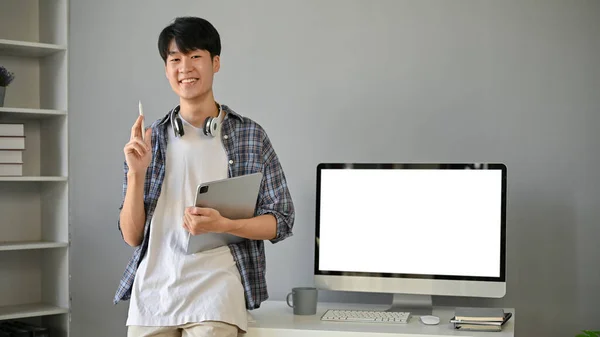 フランネルのシャツを着たハンサムなアジア系の若者が 現代のオフィスにタブレットを手にして立っている テーブルの上にある白いスクリーンのモックアップは — ストック写真