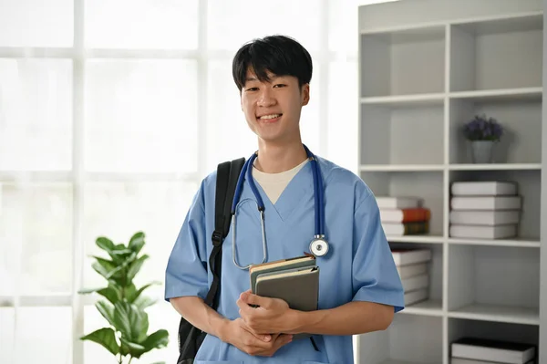 A portrait of a smiling and kind young Asian male doctor or medical student in scrubs stands in the office.