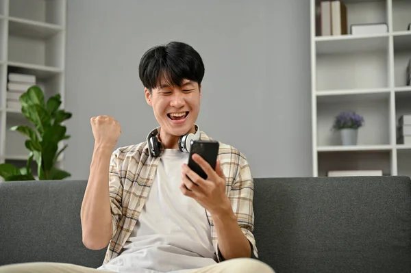 一位身穿休闲装的快乐而微笑的亚洲年轻人 坐在客厅的沙发上 看着他的智能手机屏幕 举手庆祝他的好消息 — 图库照片