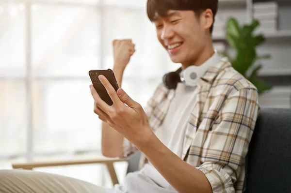 在特写中 一位兴高采烈 快乐的亚洲年轻人俯瞰着智能手机屏幕 坐在客厅的沙发上庆祝他的好消息 — 图库照片