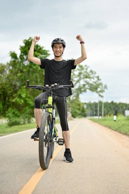 Mutlu ve sağlıklı, spor giyimli formda genç bir Asyalı ve bisiklet kaskı bisikletine binmeye ya da bisiklet hedefine ulaşmaya hazır bir şekilde ellerini kaldırıyor. Neşelen ya da jestini kutla