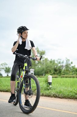 Spor kıyafetleri içinde yorgun ve terli bir Asyalı adamın portresi ve bisiklet kaskı kırsal yollarda bisiklet sürerken boynunu havluyla siliyor..