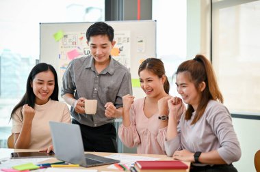 Bir grup neşeli Asyalı teknoloji geliştirici dizüstü bilgisayara bakıyor ve yeni projelerinin başarısını toplantıda kutluyorlar. takım çalışması konsepti