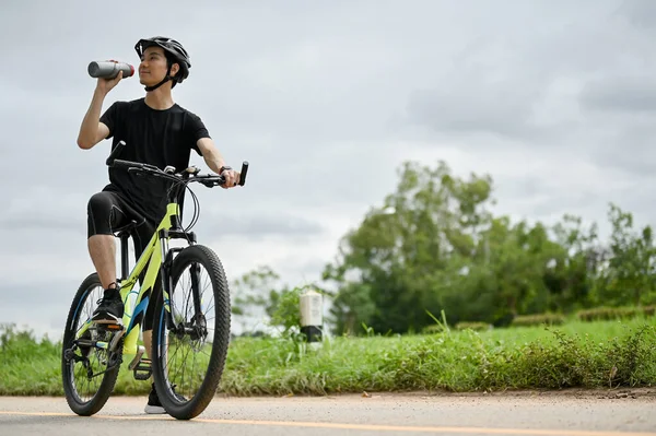 一名身穿运动服 头戴自行车头盔的健康的亚洲男子骑自行车时正在喝瓶子里的水 暑期活动概念 — 图库照片