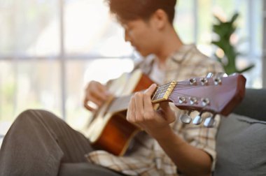 Oturma odasındaki bir kanepede gitar çalan rahat ve odaklanmış genç bir Asyalı 'nın yakın plan görüntüsü. Boş zaman ve hobi konsepti