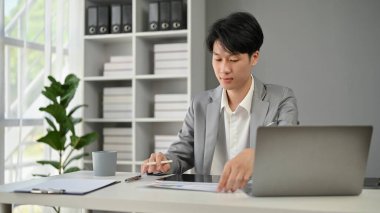Resmi bir iş elbisesi içinde profesyonel ve zeki bir Asyalı işadamı ofisindeki masasında iş belgelerini okumaya odaklanır..