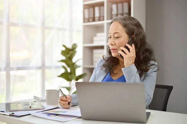 stock image A professional mature Asian female boss is on the phone with a supplier to deal a new contract while sitting at her desk in her private office.